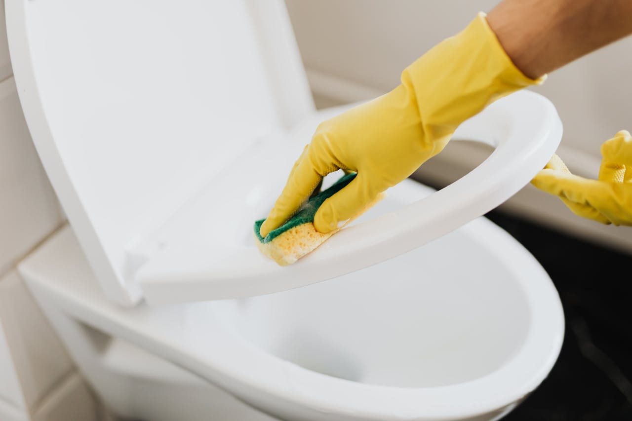 heros-img A person using yellow gloves and a sponge to clean a toilet seat indoors.